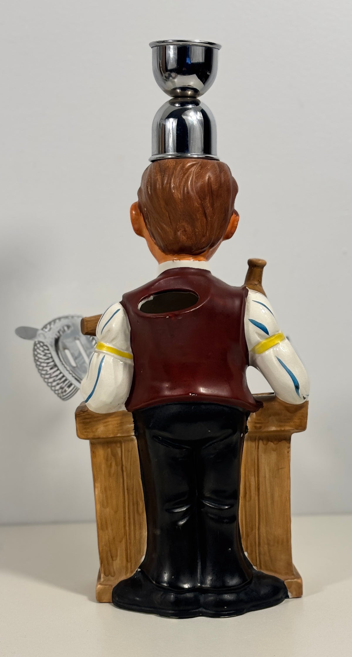 A ceramic figurine of a bartender seen from the back, balancing a metal cup on his head while holding a cocktail shaker and a bottle on a stool. The figurine features brown hair, a maroon vest, white sleeves with yellow stripes, and black pants, set against a plain light gray background.