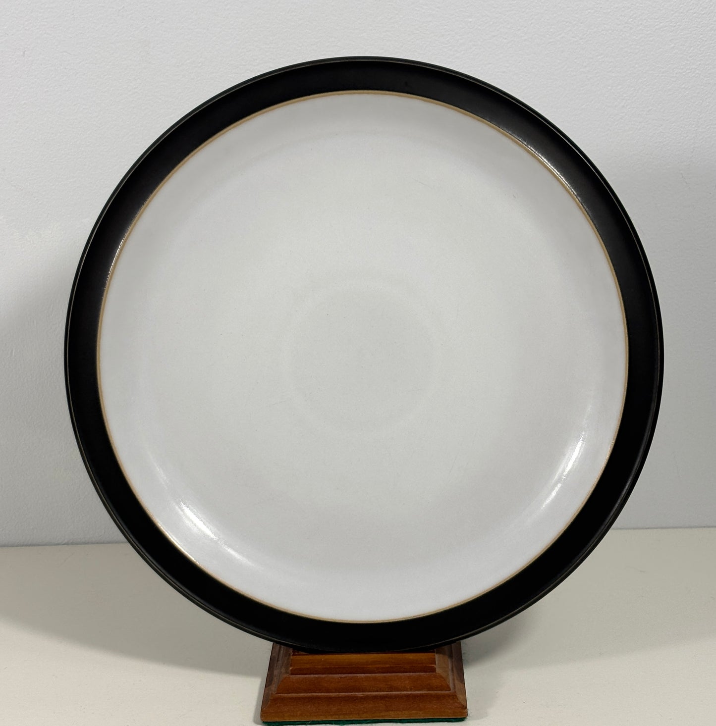 A round ceramic plate with a wide black rim and a glossy white center, displayed on a small wooden stand against a plain light background.