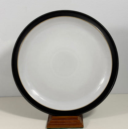A round ceramic plate with a wide black rim and a glossy white center, displayed on a small wooden stand against a plain light background.