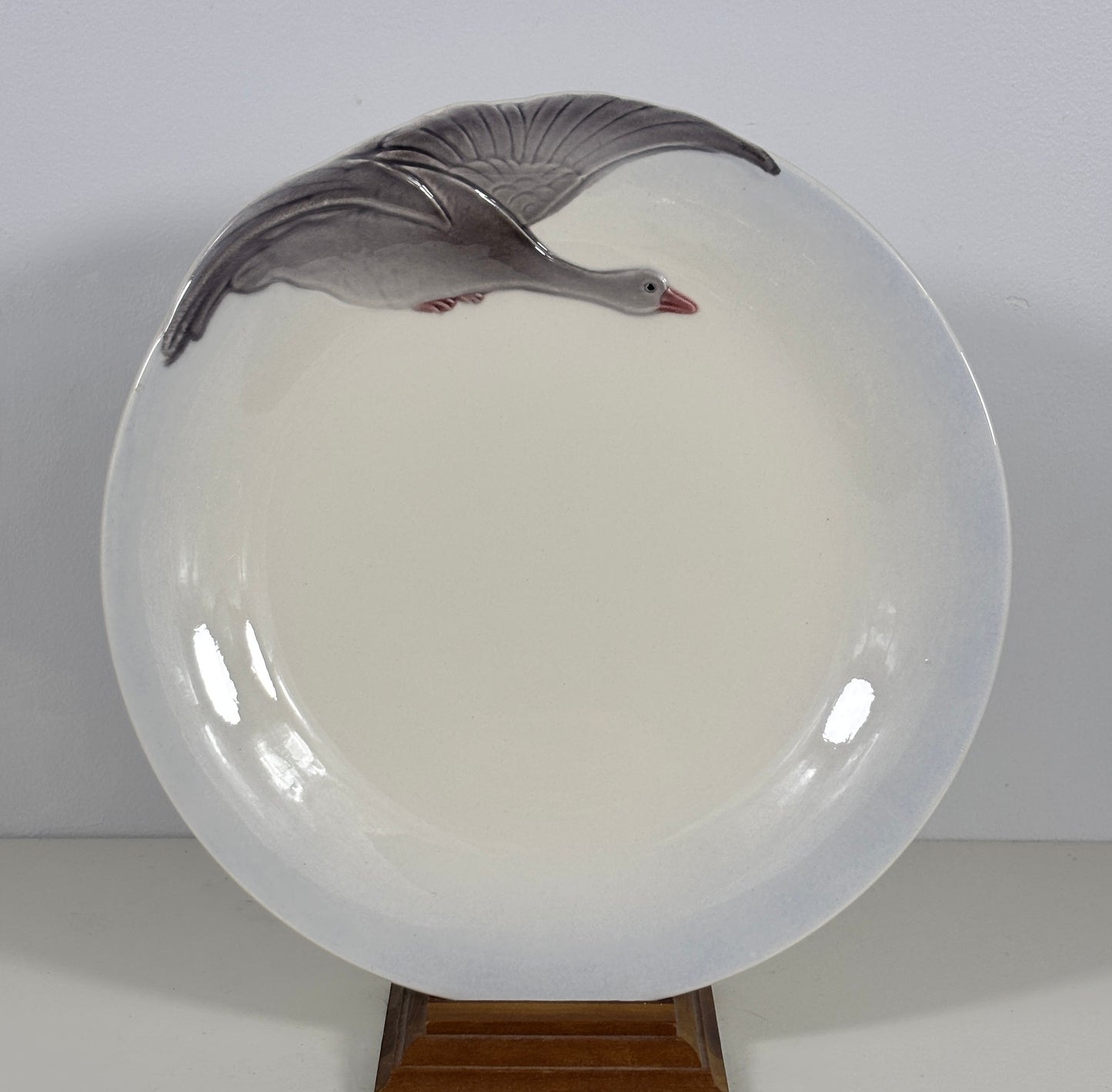 A round glossy white ceramic plate featuring a painted gray goose in flight near the top edge, displayed on a small wooden stand against a plain light background.