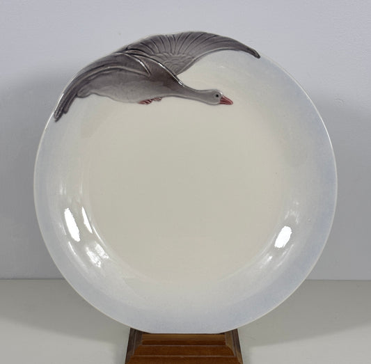A round glossy white ceramic plate featuring a painted gray goose in flight near the top edge, displayed on a small wooden stand against a plain light background.