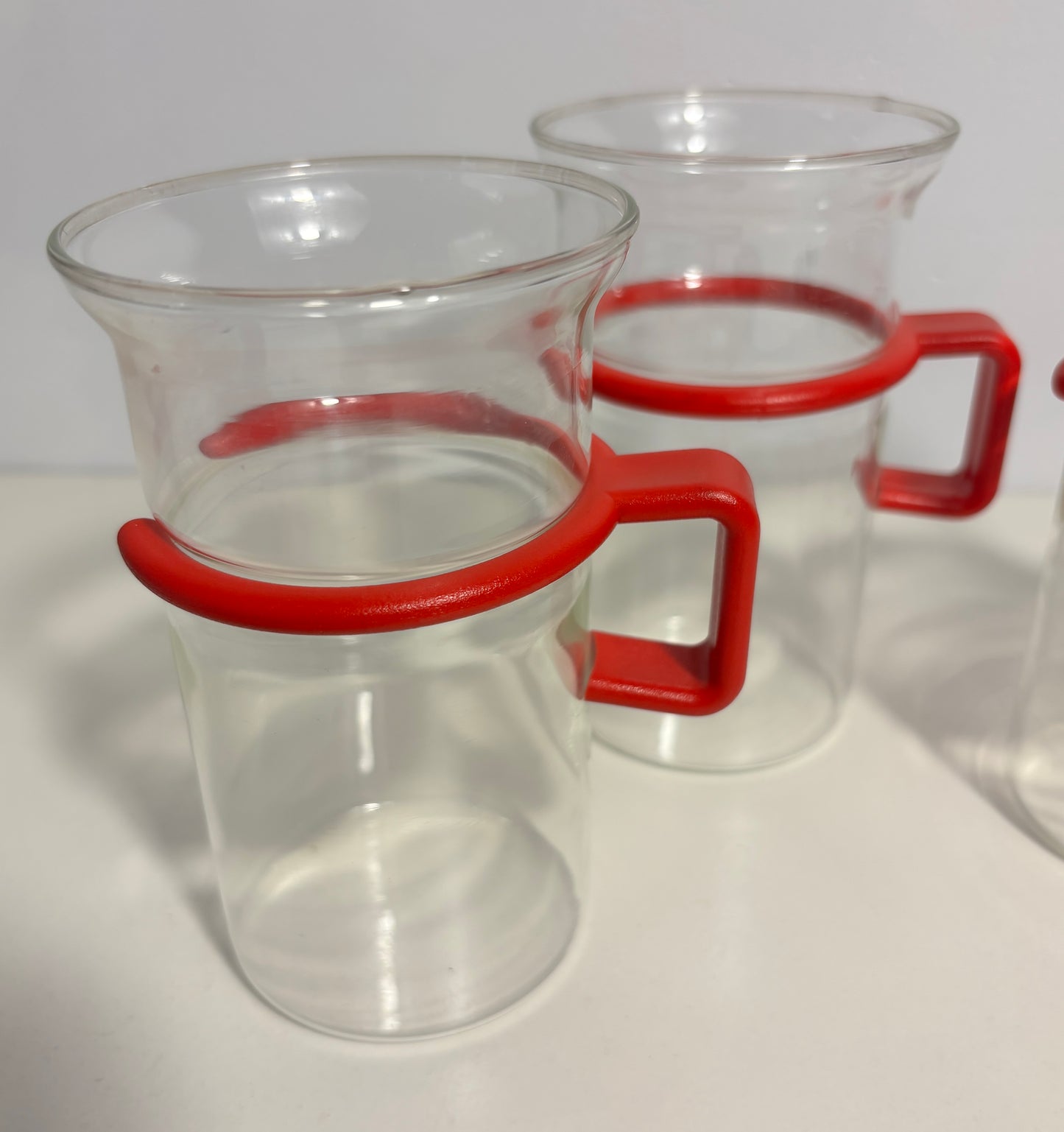 Clear glass beakers with bright red silicone handle grips, shown on a white surface with a white background.