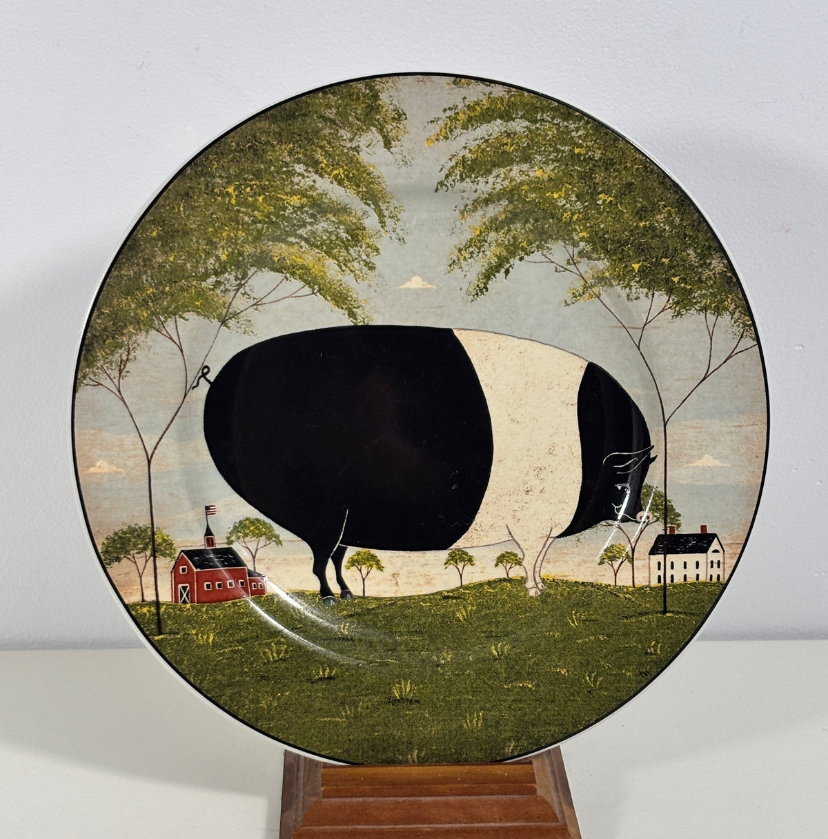 A decorative ceramic plate featuring a whimsical landscape painting of a black-and-white cow watering a red barn and small house, displayed on a wooden stand against a light gray wall.