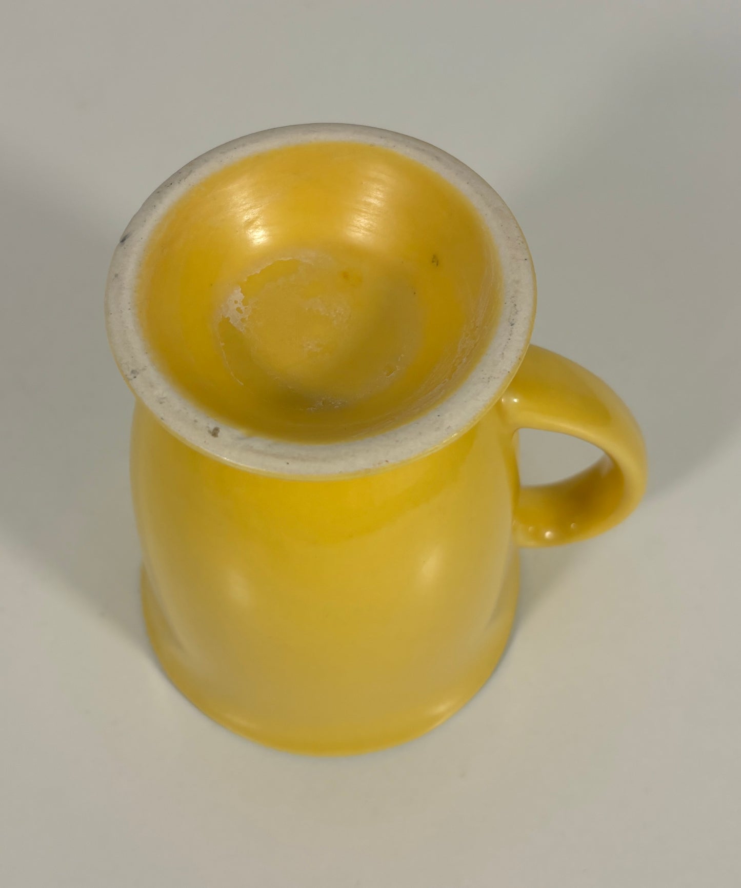 A small yellow ceramic mug with a glossy finish, featuring a curved handle and a light-colored unglazed rim, shown on a neutral light gray background.