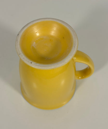 A small yellow ceramic mug with a glossy finish, featuring a curved handle and a light-colored unglazed rim, shown on a neutral light gray background.