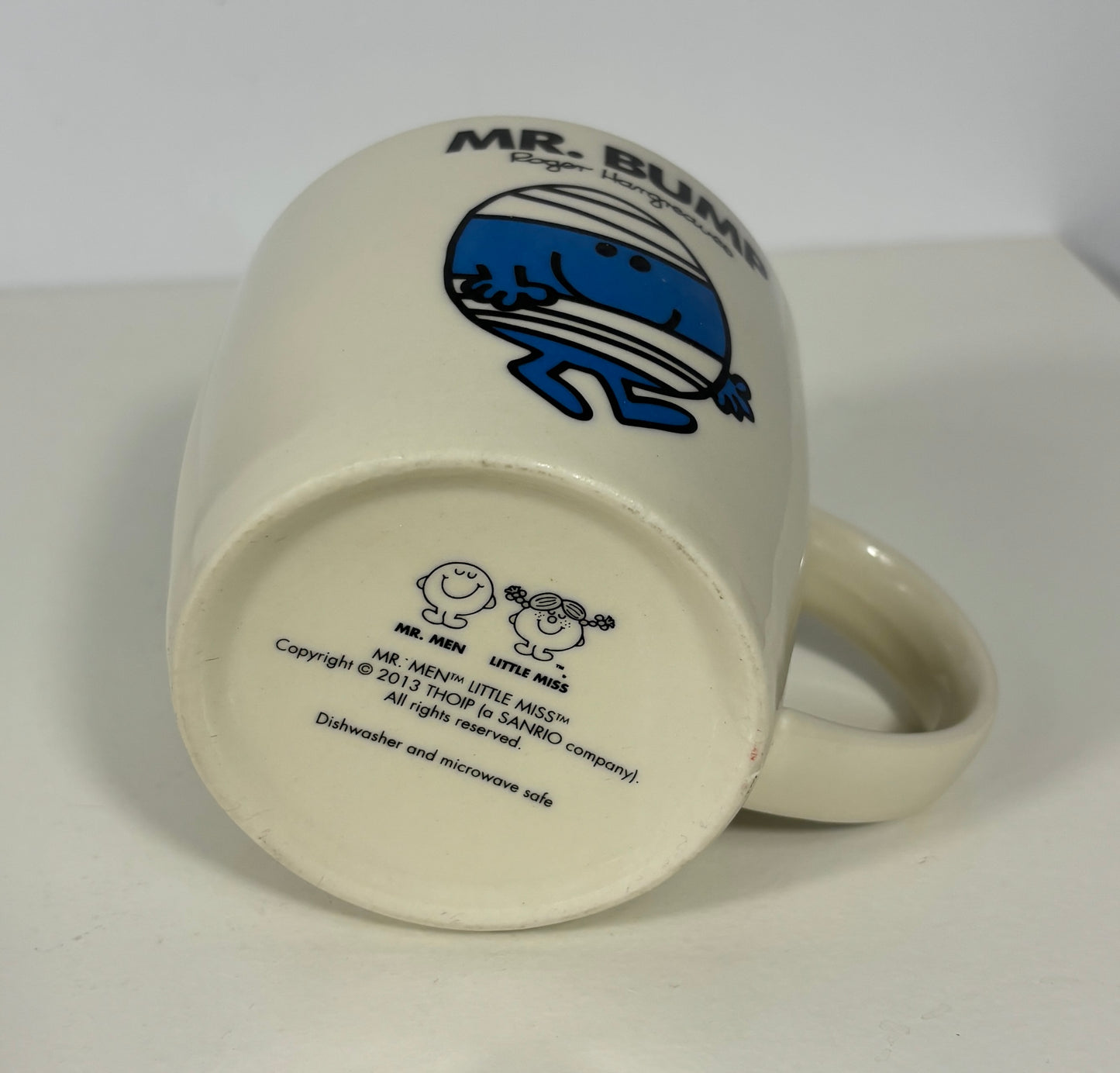 An off-white ceramic mug shown upside down, displaying the bottom side with the blue and black Mr. Bumble character graphic and printed text.
