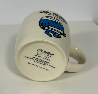 An off-white ceramic mug shown upside down, displaying the bottom side with the blue and black Mr. Bumble character graphic and printed text.