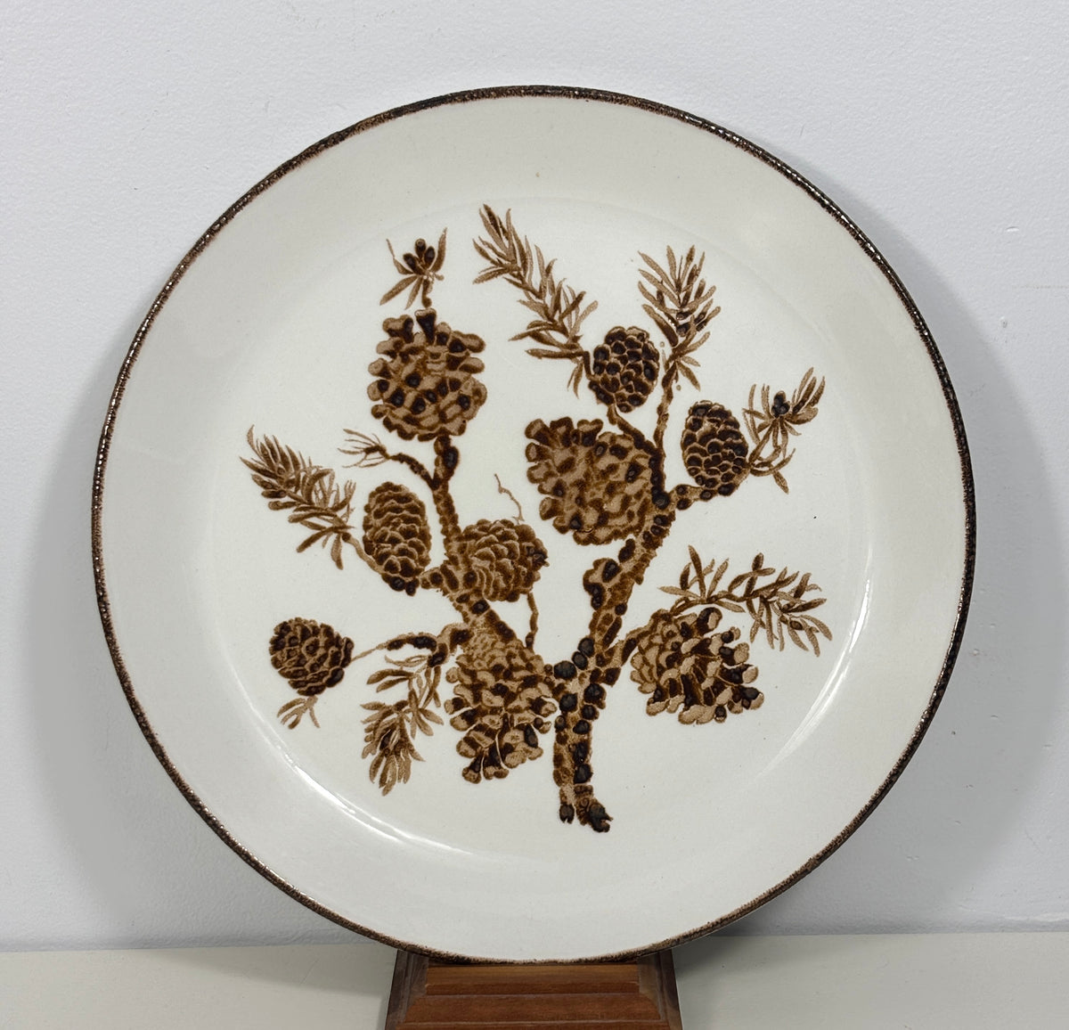 A round ceramic plate with a white glaze, featuring a brown painted pine branch design with pinecones and needles, and a rustic dark brown rim, displayed on a wooden stand against a white wall.