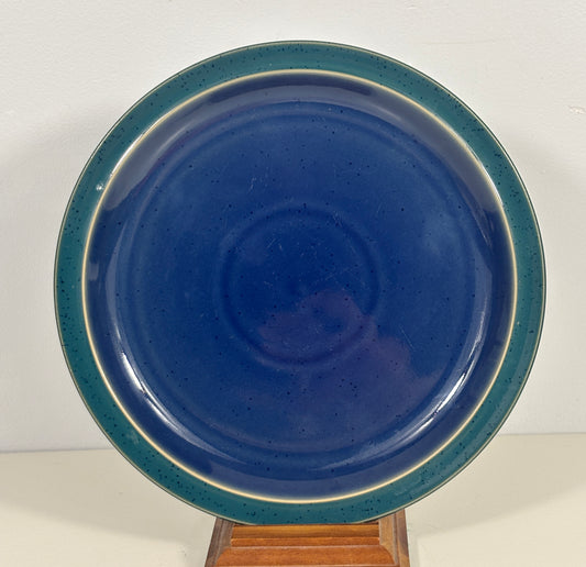 A round ceramic plate with a glossy deep blue center and a speckled teal-green rim, displayed on a small wooden stand against a plain white background.