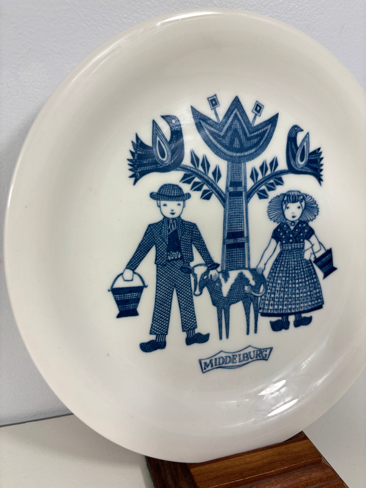 A white ceramic plate with a blue folk-art style illustration of a woman and child, photographed on a light surface with part of a wooden object visible at the right edge.