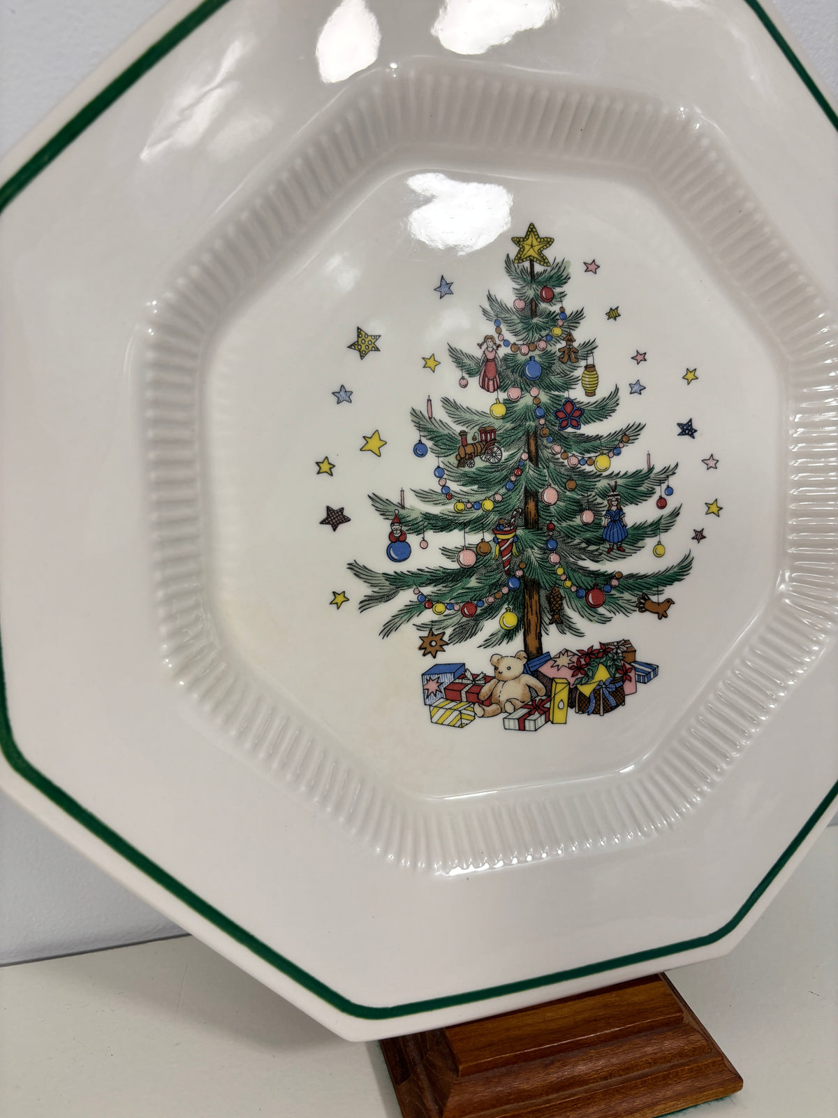 A white octagonal ceramic plate with a green rim, featuring a printed Christmas tree illustration in the center surrounded by stars and wrapped gifts, displayed on a wooden stand.