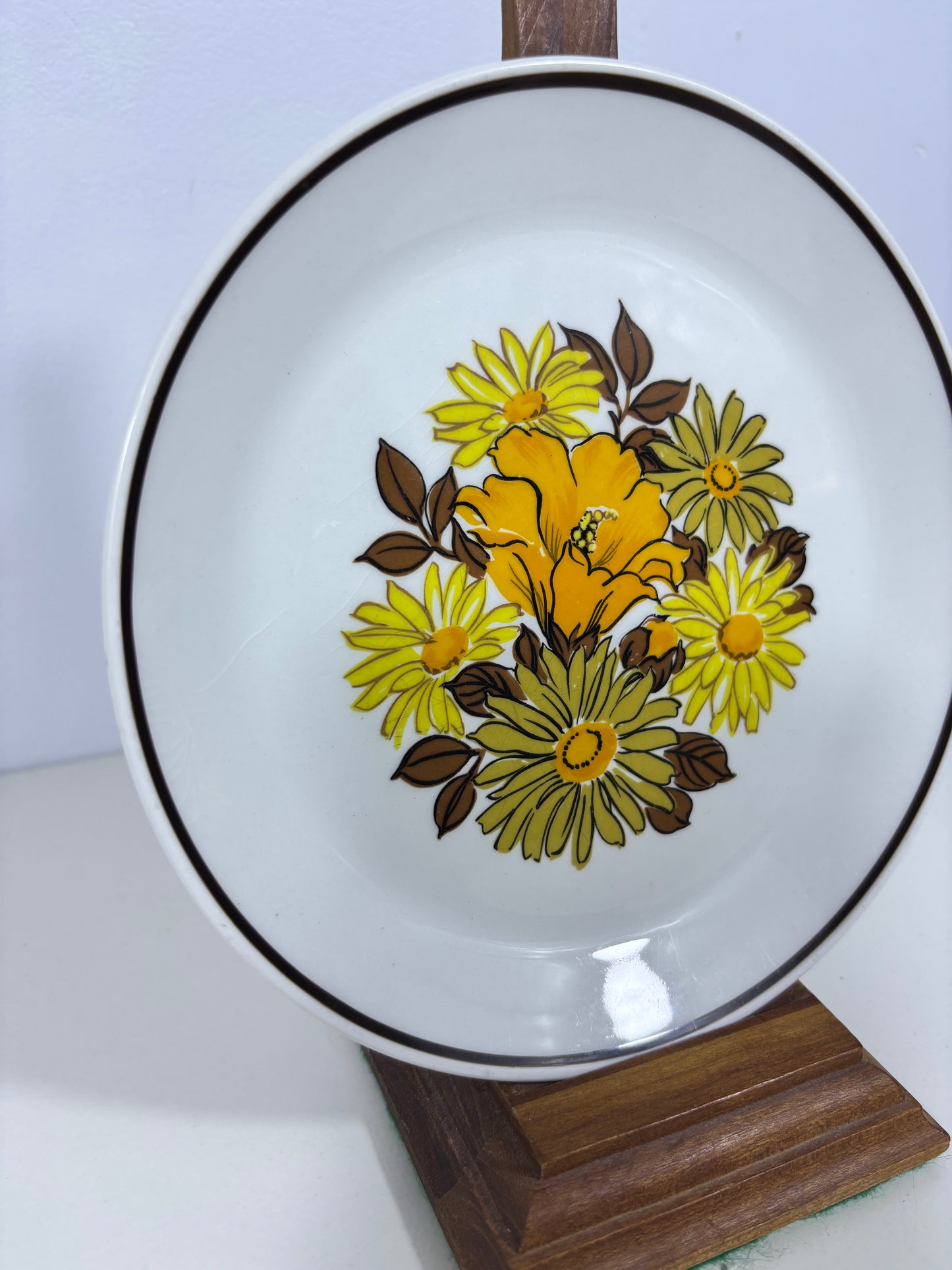 A white ceramic plate with a floral pattern featuring yellow and green flowers with brown leaves, accented by a thin black rim, shown on a white background with a wooden block.