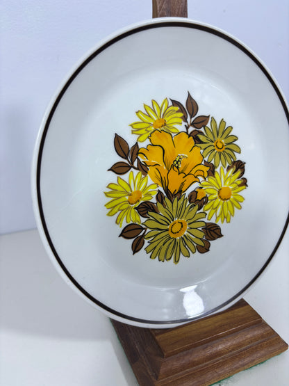 A white ceramic plate with a floral pattern featuring yellow and green flowers with brown leaves, accented by a thin black rim, shown on a white background with a wooden block.