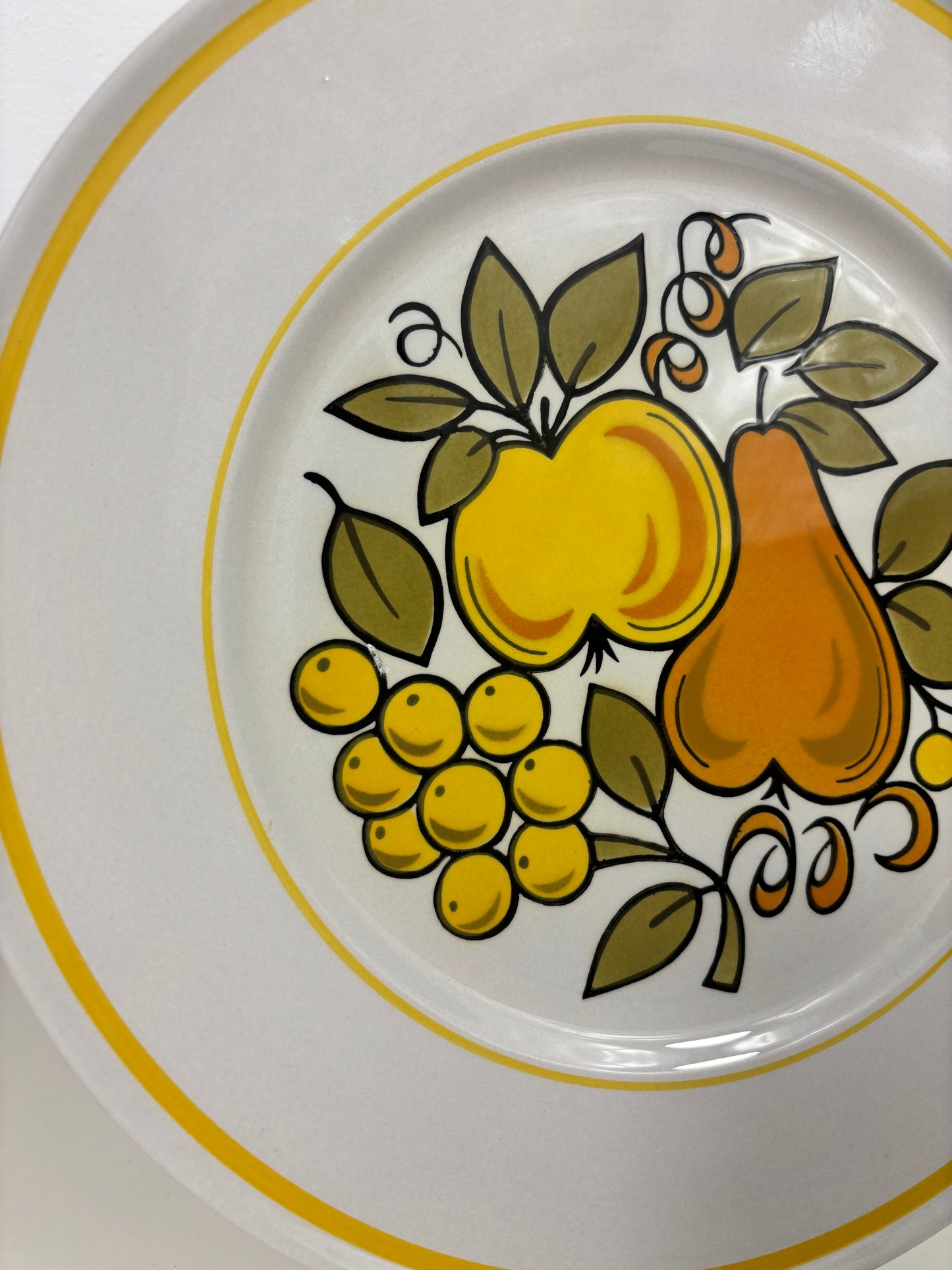 A close-up of a white ceramic plate featuring a vintage fruit pattern with a yellow apple, orange pear, yellow grapes, and green leaves, accented by yellow stripes on the rim.