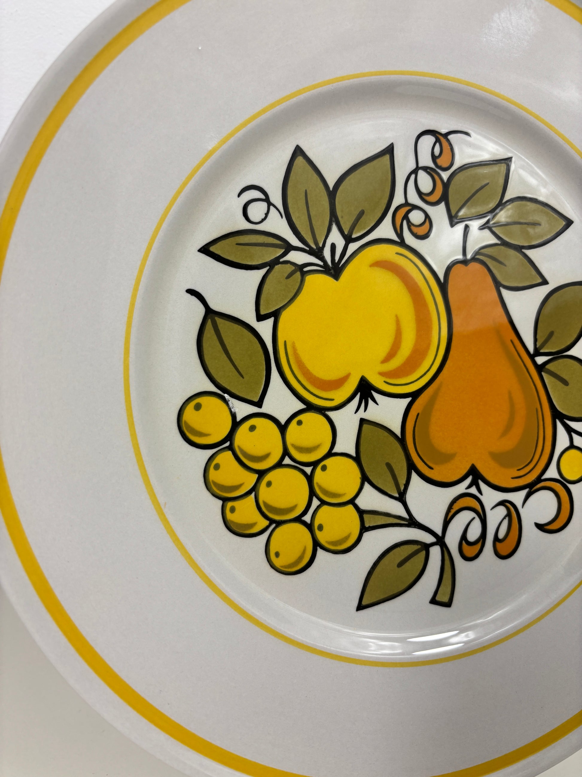 A close-up of a white ceramic plate featuring a vintage fruit pattern with a yellow apple, orange pear, yellow grapes, and green leaves, accented by yellow stripes on the rim.