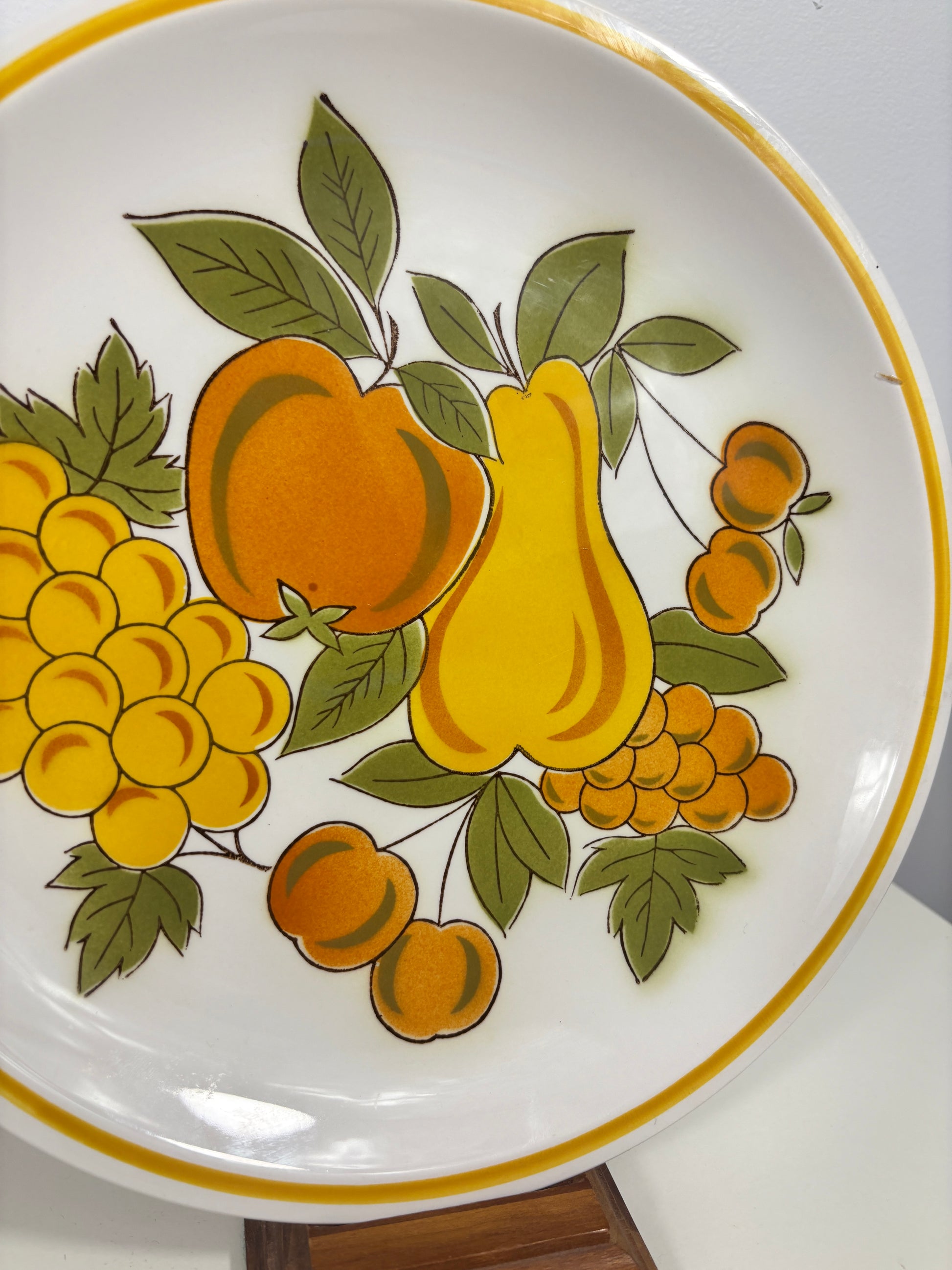 A white ceramic plate featuring a retro-style fruit pattern with yellow pears, orange peaches, green leaves, and yellow grapes, finished with a yellow-gold rim.