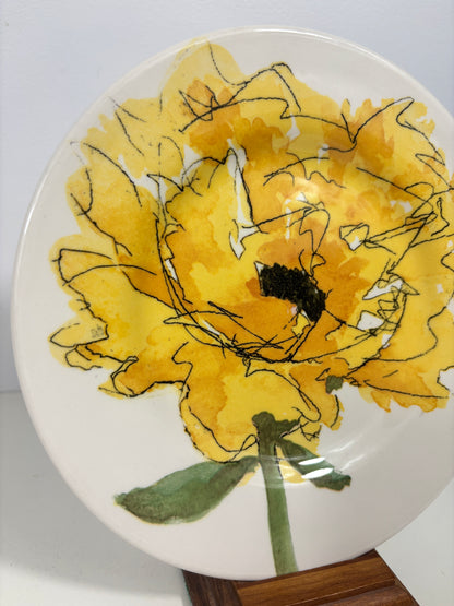 A white ceramic plate featuring a hand-painted yellow flower with black sketch lines, green leaves, and stem, displayed on a wooden stand against a white background.