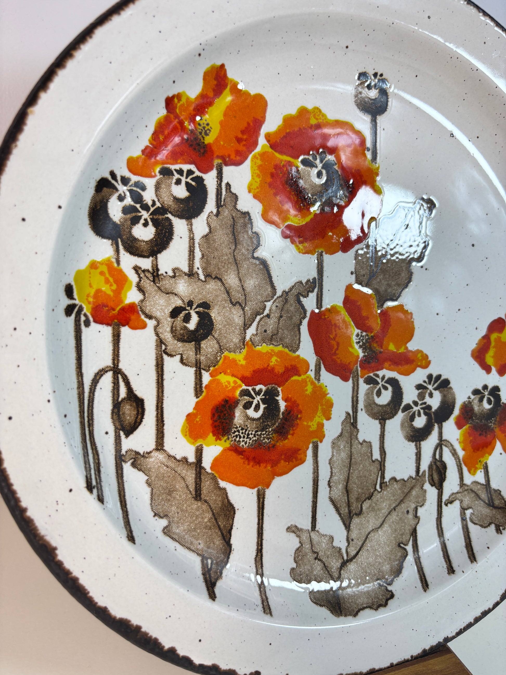 A close-up of a ceramic plate with a hand-painted poppy floral design in orange, red, yellow, and brown on a white background.
