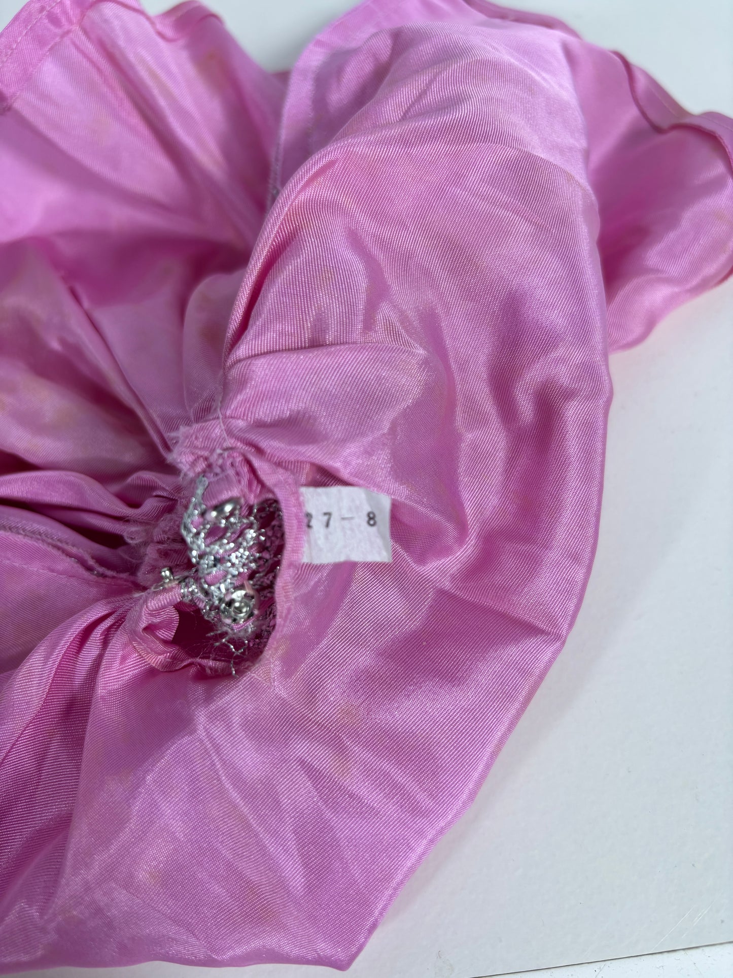 A close-up of a pink sheer fabric accessory, likely a hair scrunchie or gathered fabric piece, featuring a silver rhinestone embellishment and a small white size tag reading '27-8'.