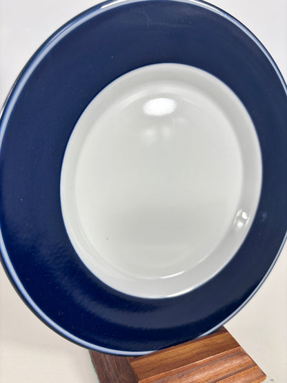 A ceramic plate with a wide navy blue rim and a glossy white center, displayed on a wooden stand against a light background.