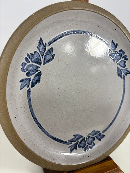 A ceramic plate with a speckled off-white interior, decorated with a blue floral pattern arranged in an oval border, and a rustic brown exterior, displayed on a wooden stand.