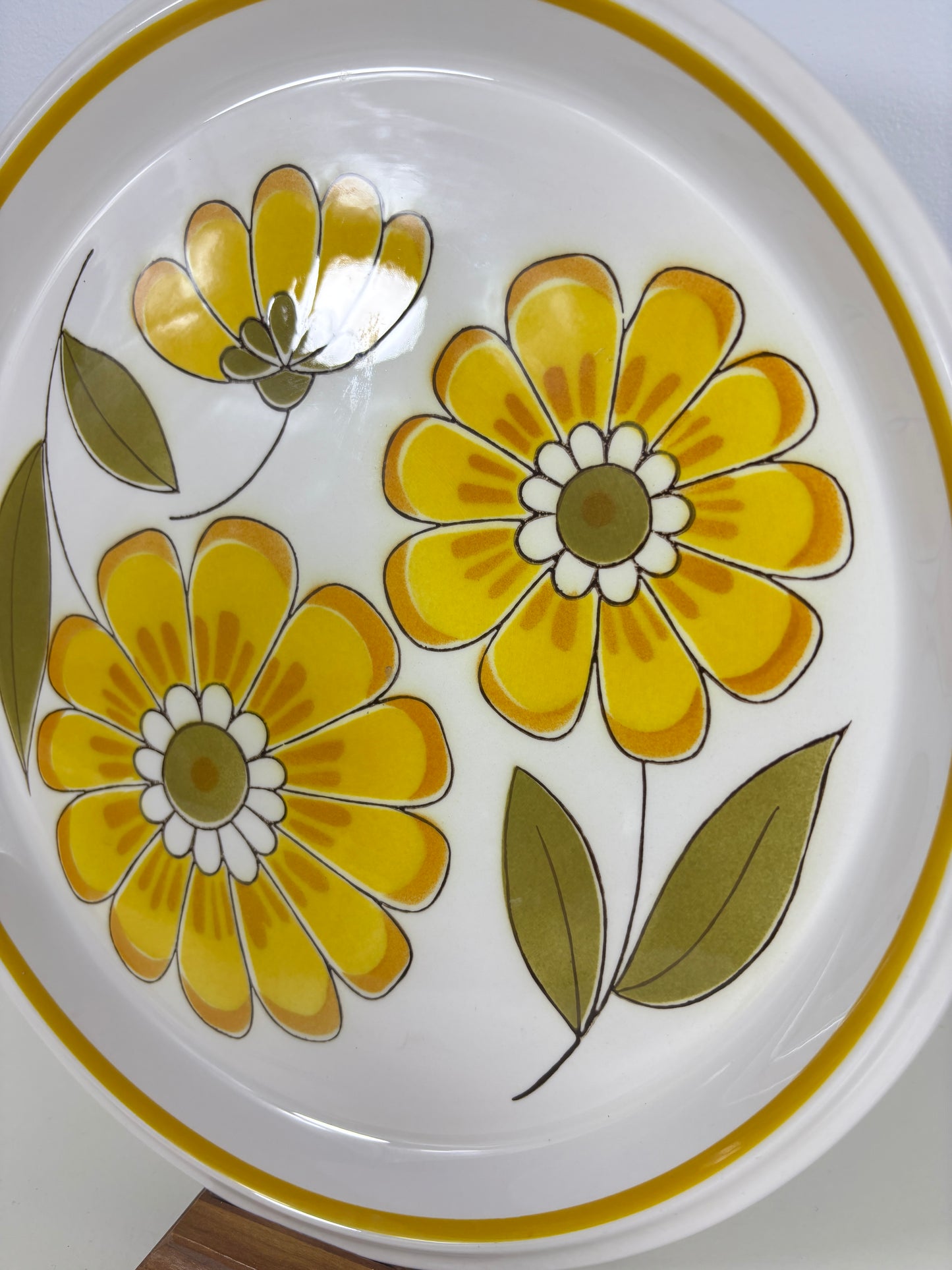 A close-up of a white ceramic plate featuring a retro floral pattern with yellow flowers, white inner petals, and olive green leaves, finished with a gold rim.