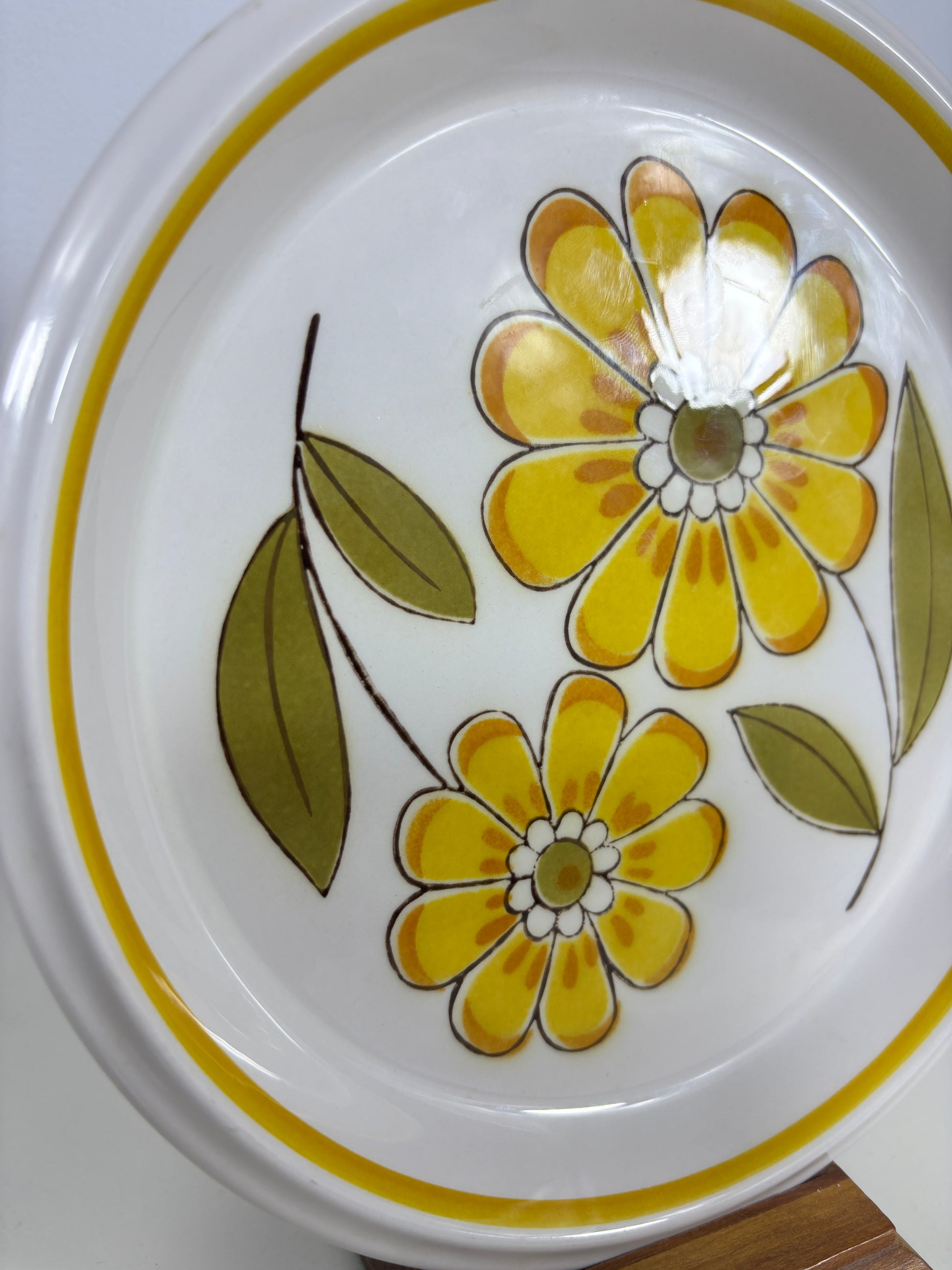 A close-up of a white ceramic plate featuring a retro floral pattern with yellow flowers and green leaves, accented by a yellow rim.