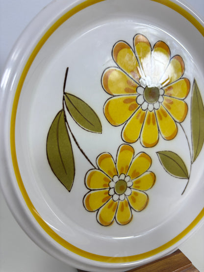 A close-up of a white ceramic plate featuring a retro floral pattern with yellow flowers and green leaves, accented by a yellow rim.