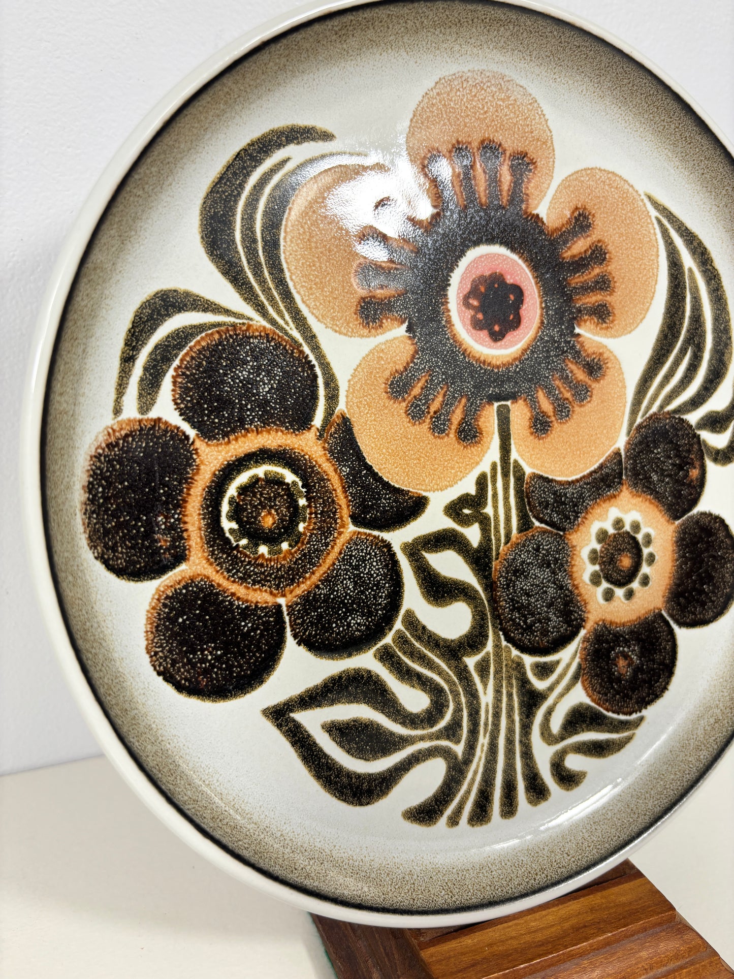 A round ceramic plate with a retro floral pattern in dark brown, beige, and black on a white glaze, displayed on a wooden stand.