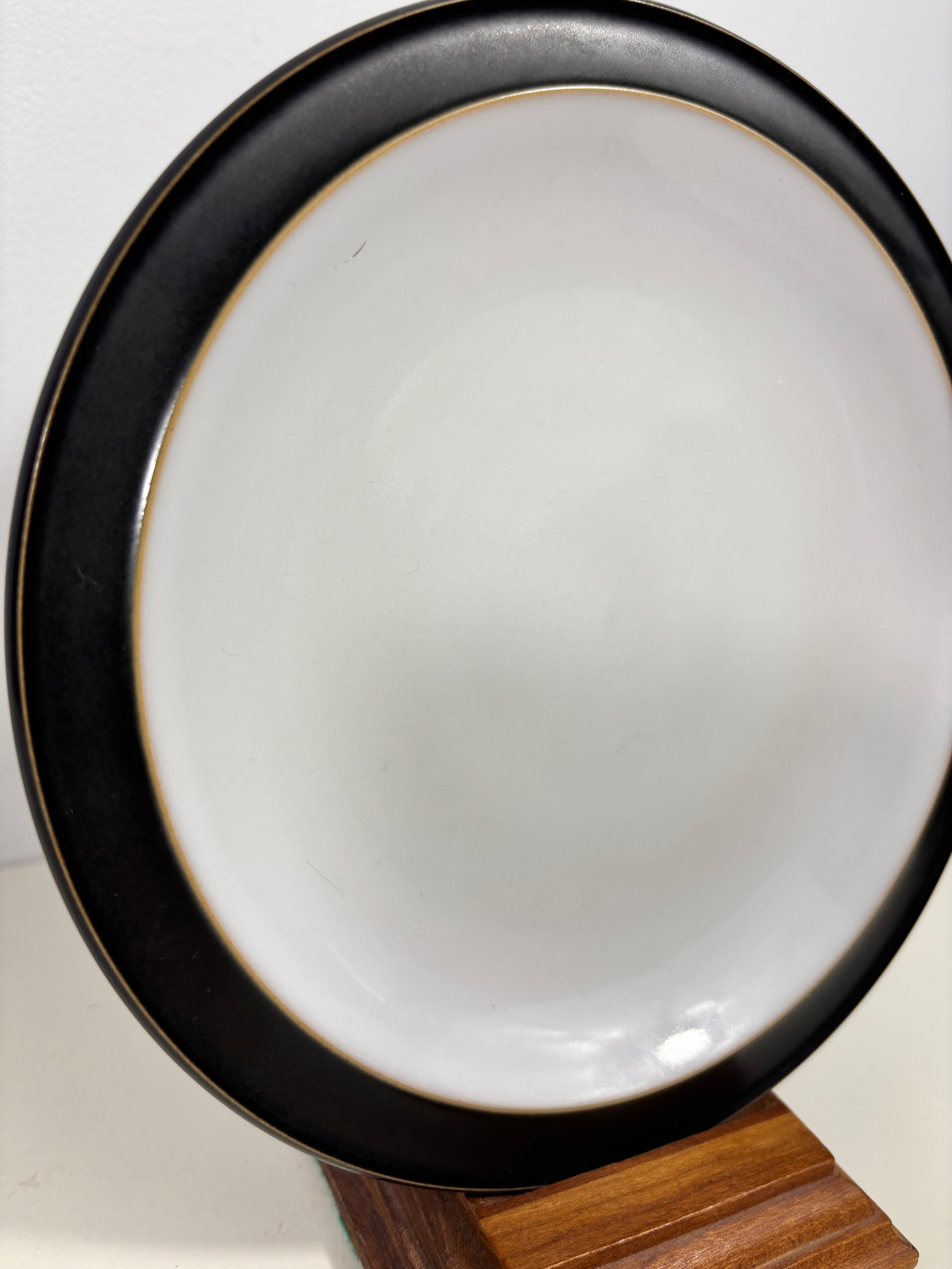A round ceramic plate with a glossy white interior and a wide black rim accented by a thin gold line, displayed on a wooden stand against a plain white background.