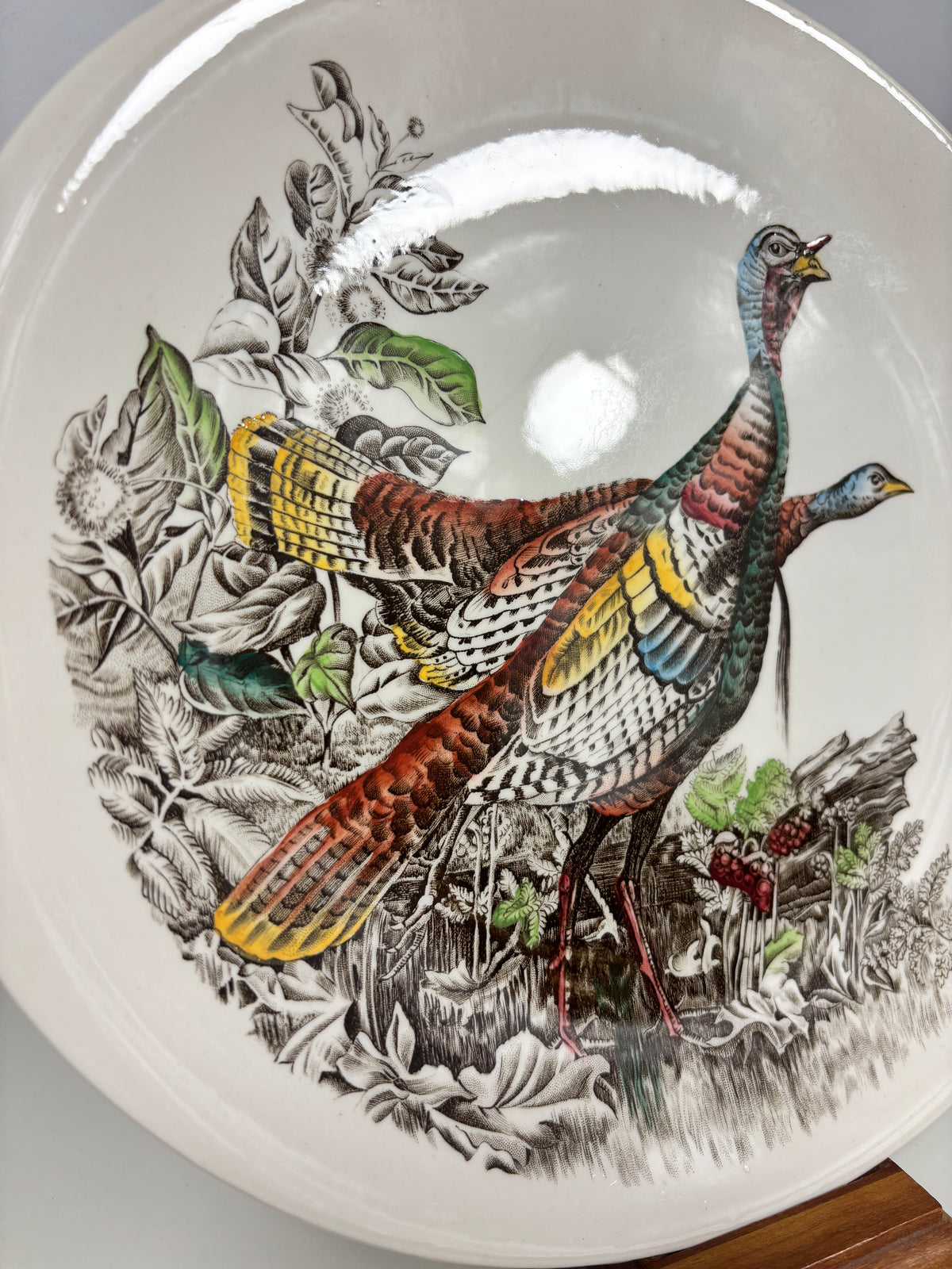 A close-up of a ceramic plate featuring a detailed illustration of two wild turkeys in a natural forest setting with leaves and berries. The plate has a glossy white glaze with a prominent overhead light reflection.