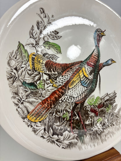 A close-up of a ceramic plate featuring a detailed illustration of two wild turkeys in a natural forest setting with leaves and berries. The plate has a glossy white glaze with a prominent overhead light reflection.