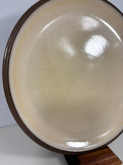 Close-up of a round ceramic plate or dish with a glossy cream-beige interior and a dark brown rim, displayed on a wooden stand against a light background.