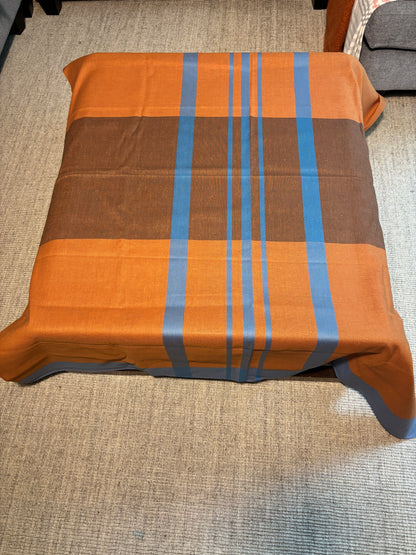 A square table covered with a plaid tablecloth in orange, brown, and blue, shown from a top-down view on a textured beige carpet.