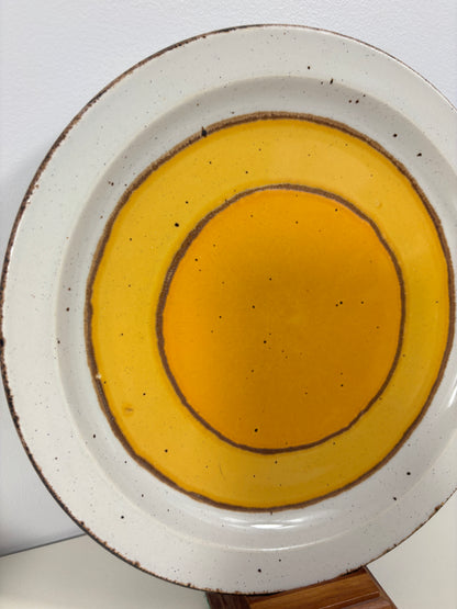 A top-down view of a ceramic plate with concentric circular patterns in yellow and orange glazes, featuring a speckled off-white body and a rustic brown rim.