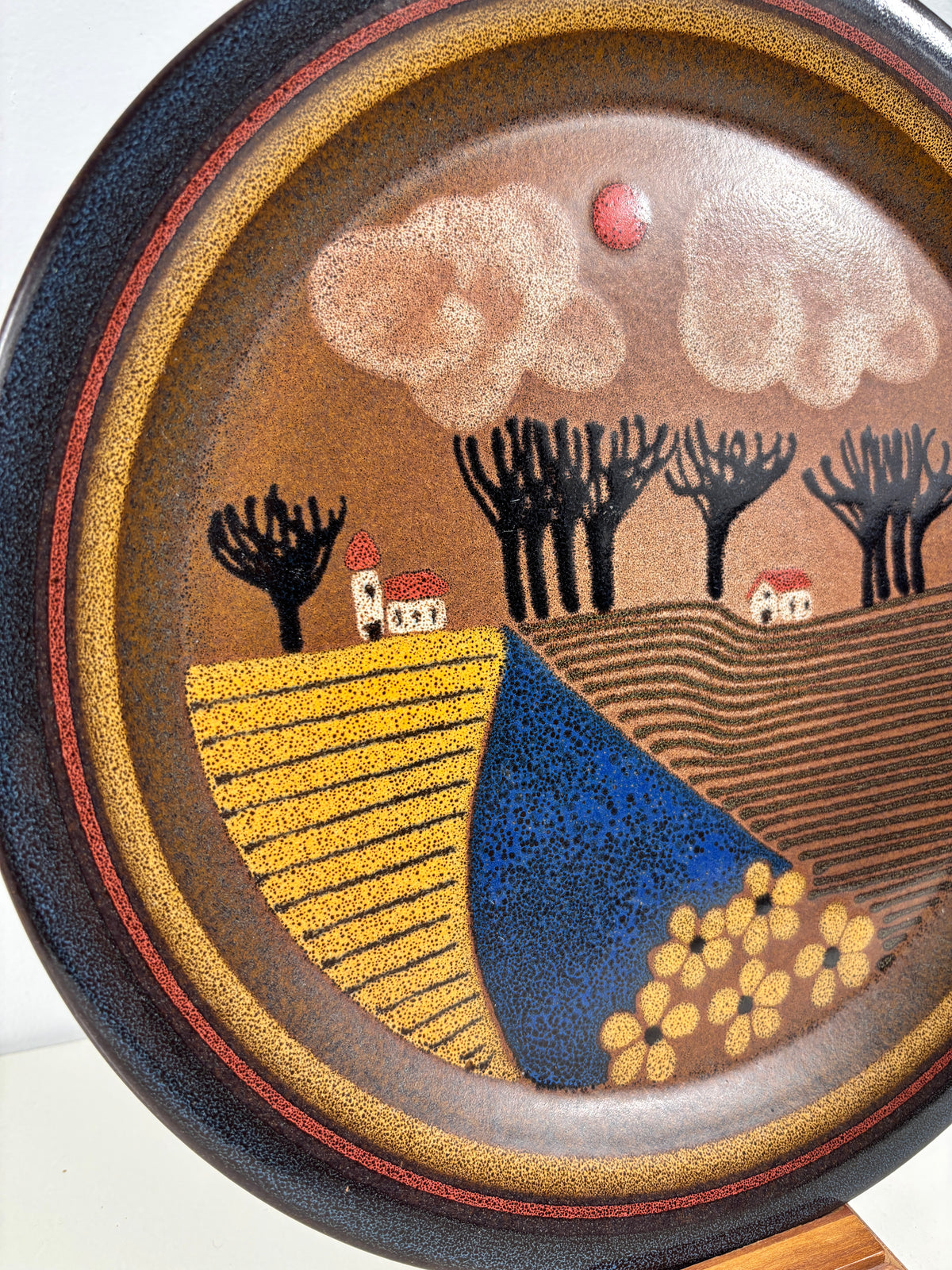 A close-up of a round ceramic plate or decorative dish featuring a stylized landscape scene with yellow fields, black trees, small houses, a blue foreground shape, and a brown sky with cream clouds.