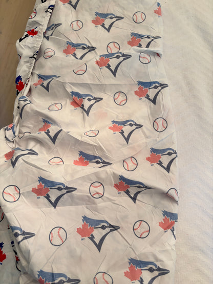 A white fitted sheet or duvet cover featuring an all-over repeating print of the Toronto Blue Jays baseball team logo, including blue jay heads, baseballs, and red maple leaves.