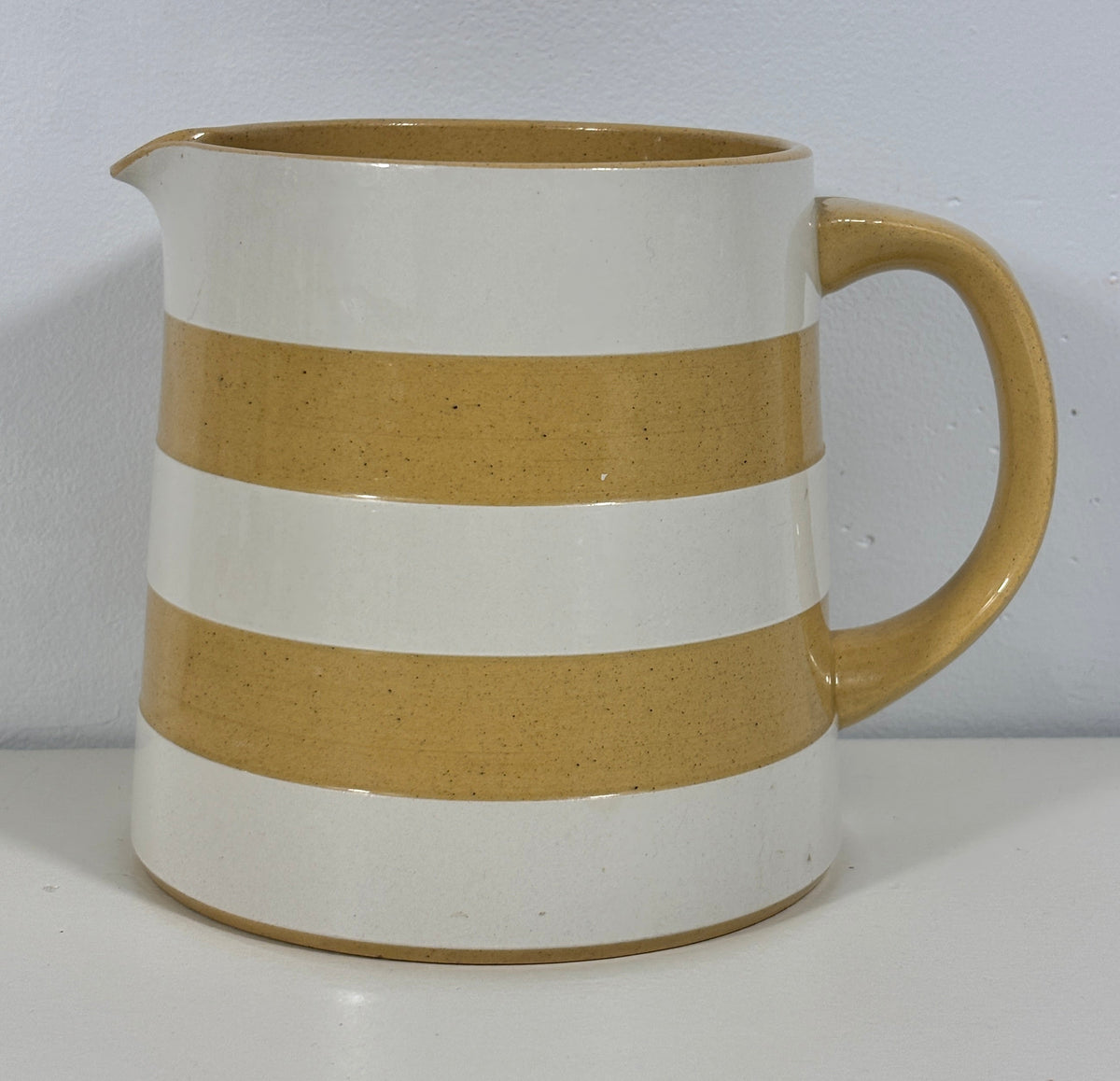 A ceramic pitcher with a glossy finish, featuring alternating horizontal stripes of white and a warm sandy beige, with a matching beige handle and spout.