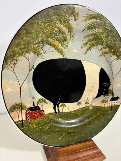 An oval decorative plate featuring a whimsical folk-art style scene with a large black-and-white cow, green trees, a red barn, and a house, displayed on a wooden stand against a light background.