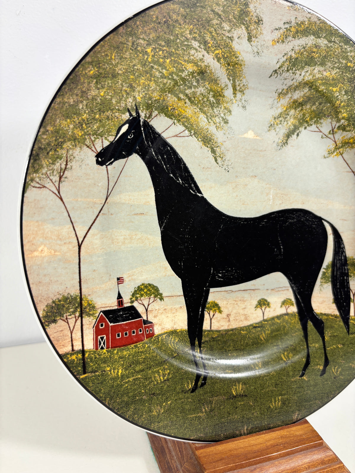 A round decorative plate featuring a hand-painted barn scene with a large black horse, green trees, and a red barn, displayed on a wooden stand.