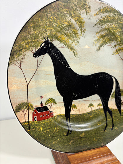 A round decorative plate featuring a hand-painted barn scene with a large black horse, green trees, and a red barn, displayed on a wooden stand.