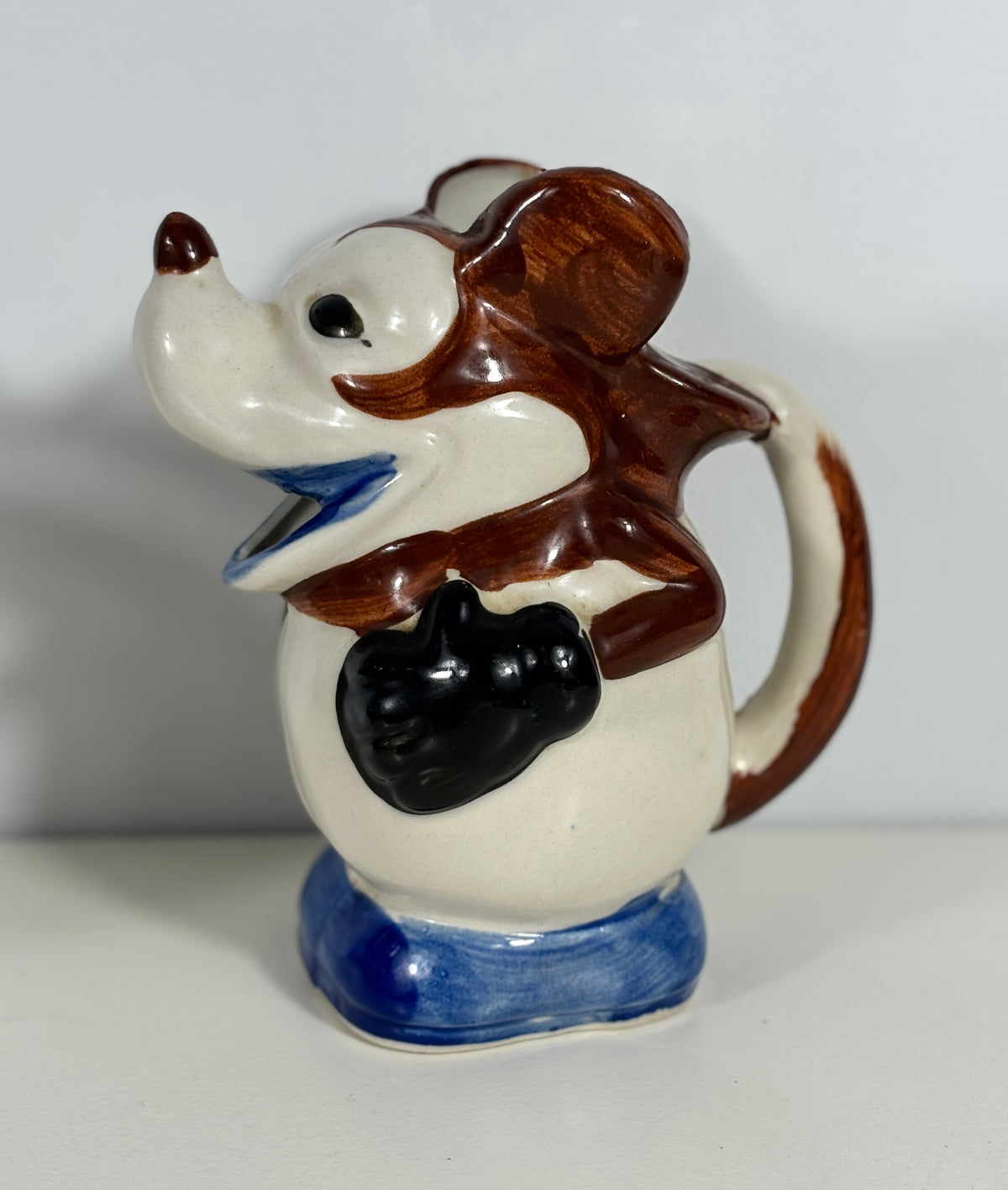 A ceramic pitcher shaped like a cartoon mouse, featuring a cream-colored body, brown ears and tail, blue shoes, and a black patch on the chest, set against a plain light gray background.
