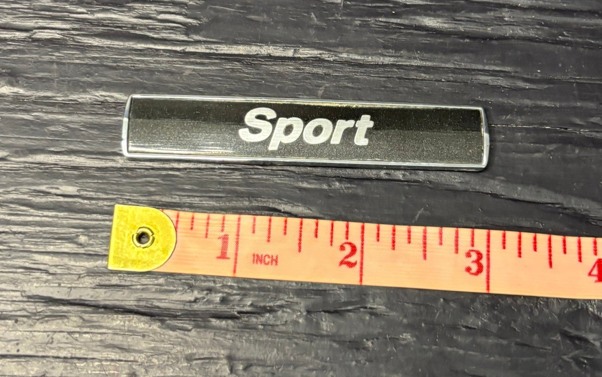 Metallic 'Sport' badge with a ruler for scale on a dark wooden surface