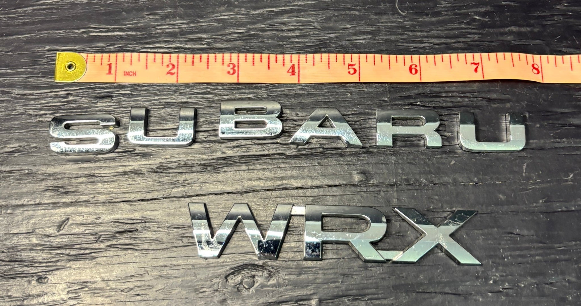 Metallic 'SUBARU' and 'WRX' emblems with a ruler for scale on a wooden surface.