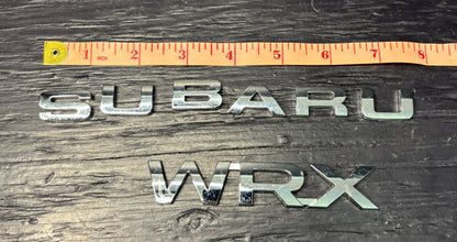 Metallic 'SUBARU' and 'WRX' emblems with a ruler for scale on a wooden surface.