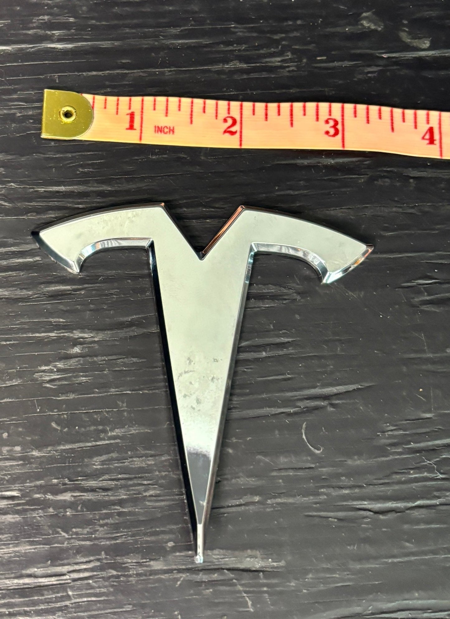 Metallic Tesla logo with a ruler for scale on a dark surface
